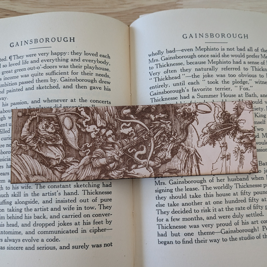 A leather bookmark with the antique art of angels and demons in battle engraved on it. The angels battle demons bookmark is resting on the pages of an open book.