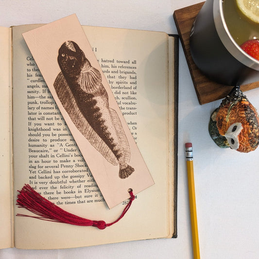 The bookmark resting on the pages of an open book. The rectangular bookmark has the antique art of a atlantic wolffish engraved on it. The bookmark also has a wine red tassel.