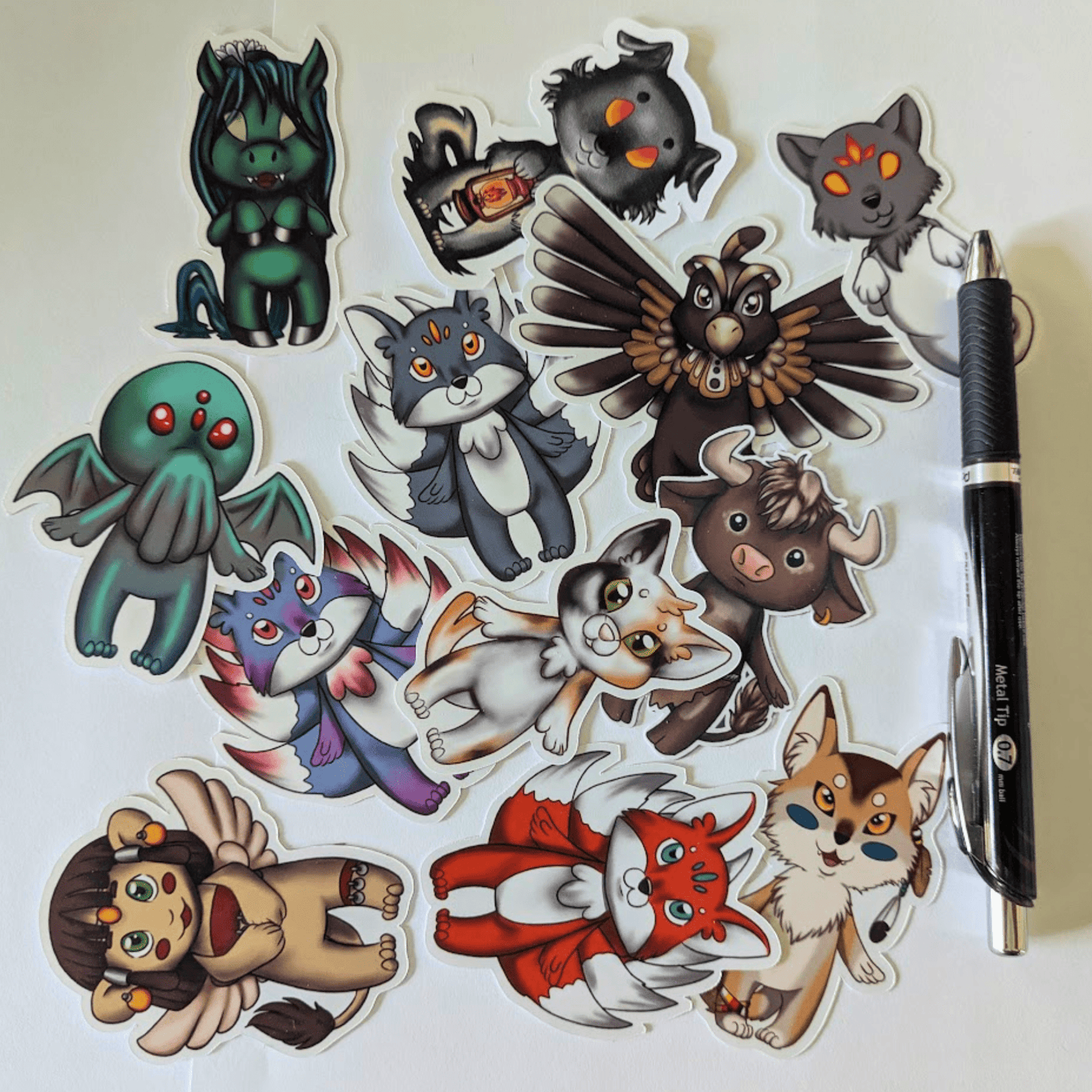 Chibi Mythos Sticker Set 1, features mythos chibis Cthulhu, kitsune, inugami, church grimm, Nekomata, spinx, Thunderbird, coyote the trickster, minotaur and Kelpie. Next to a pen for scale. Chibi Mythos Sticker Set 1 stickers Raven King Crafts