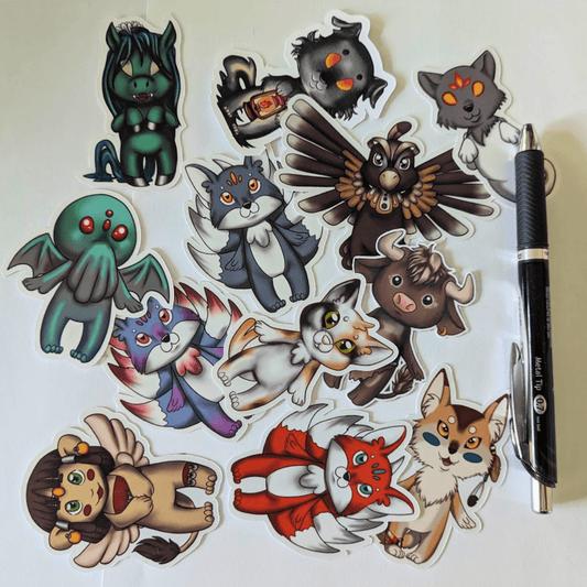 Chibi Mythos Sticker Set 1, features mythos chibis Cthulhu, kitsune, inugami, church grimm, Nekomata, spinx, Thunderbird, coyote the trickster, minotaur and Kelpie. Next to a pen for scale. Chibi Mythos Sticker Set 1 stickers Raven King Crafts