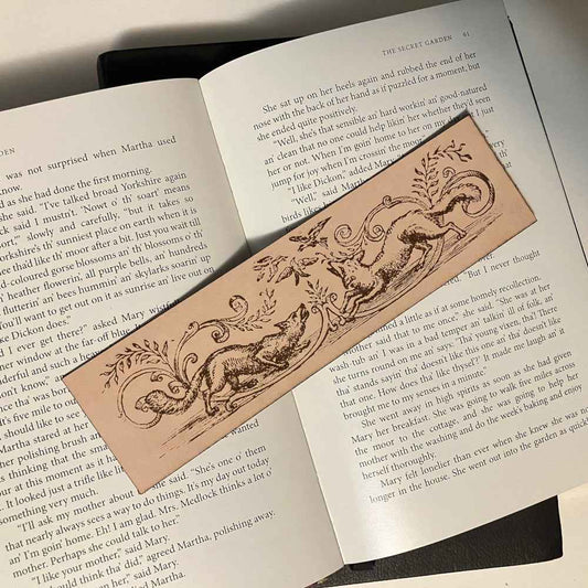 A rectangular leather bookmark with the historical art of 2 foxes trying to catch birds engraved on it. The leather fox bookmark is resting on the pages of an open book.