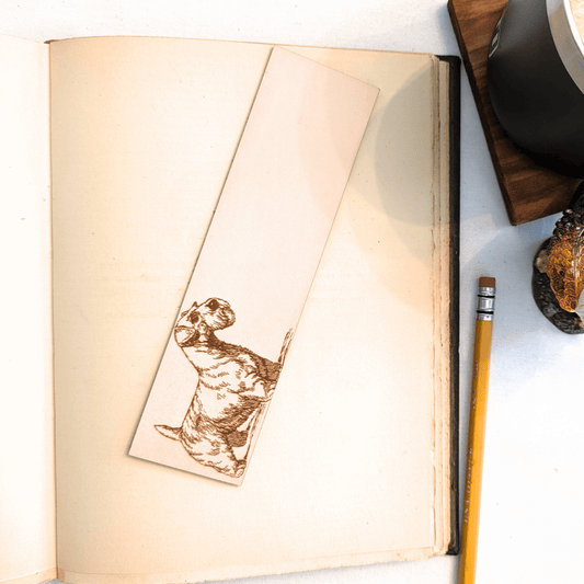 The leather Sealyham, dog bookmark resting on the pages of an open book. to the right of it is a pencil and a glass for scale. Leather Sealyham Terrier Bookmark (toy dog) Bookmarks Raven King Crafts