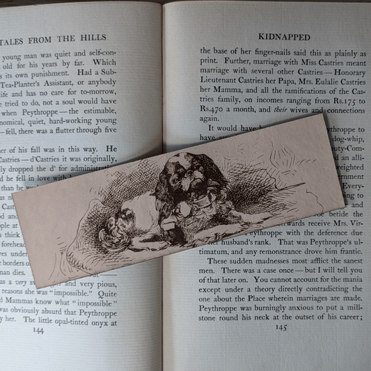 A rectangular leather bookmark with the historical art of a small spaniel dog engraved on it. The bookmark is resting on the pages of an open book. Leather Spaniel Bookmark Bookmarks Raven King Crafts