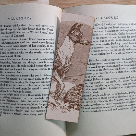 A rectangular leather bookmark with the historical art of a Whippet dog engraved on it. The bookmark is resting on the pages of an open book. Leather Whippet Dog Bookmark Bookmarks Raven King Crafts