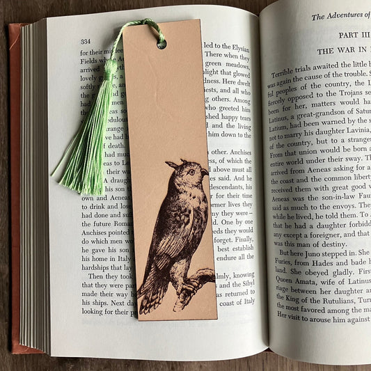 A rectangular leather bookmark with the engraving of a long eared owl on it. The owl bookmark has a green tassel. The bookmark is resting on the pages of an open book. Long-eared Owl Bookmark - Leather Bookmarks Raven King Crafts