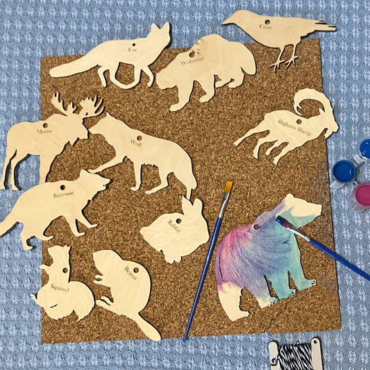 A photograph of 11 animal cutouts on a piece of cork. The bear has been painted with acrylic paints. There are 2 mini paint brushes and mini bobbin of string. North America Wildlife Ornament (Cutout) Kit Art & Craft Kits Raven King Crafts
