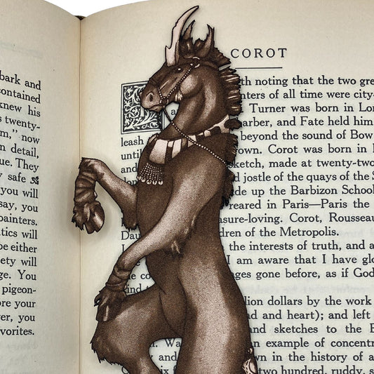 A leather bookmark shaped like a unicorn resting on the pages of an open book. The leather unicorn bookmark shows that the unicorn is wearing a collar, harness or bridle, trail cuff and leg wrappings.