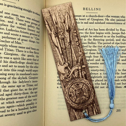 A leather bookmark with the art of a cache of weapons engraved onto it. This bookmark also has a light blue tassel attached. The bookmark is resting on the pages of an open book.