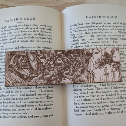 A leather bookmark with the antique art of angels and demons in battle engraved on it. The angels battle demons bookmark is resting on the pages of an open book. 