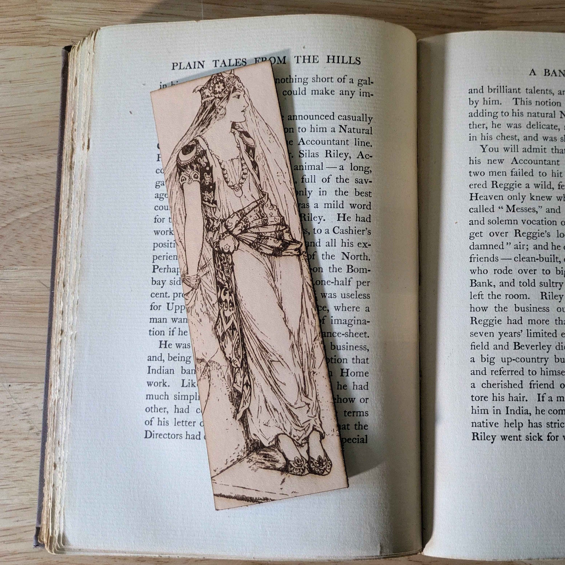 A leather rectangular bookmark with the antique art of a princess waiting engraved on it. The leather bookmark is resting on the pages of an open book.