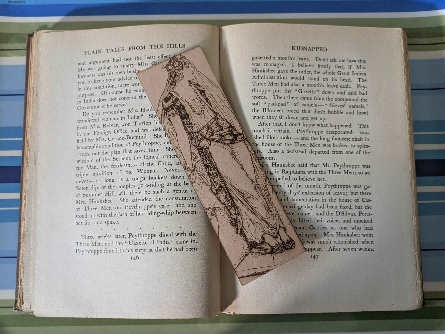 A leather rectangular bookmark with the antique art of a princess waiting engraved on it. The leather bookmark is resting on the pages of an open book.