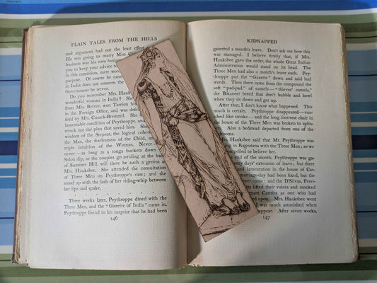 A leather rectangular bookmark with the antique art of a princess waiting engraved on it. The leather bookmark is resting on the pages of an open book.