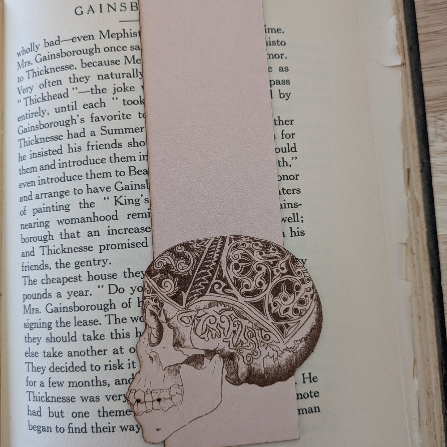 A leather bookmark engraved with the art of a skull on it. The skull bookmark is resting on the pages of an open book. 