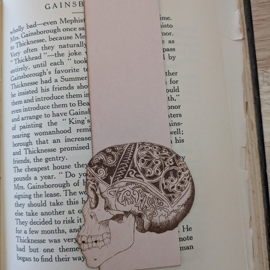 A leather bookmark engraved with the art of a skull on it. The skull bookmark is resting on the pages of an open book. 