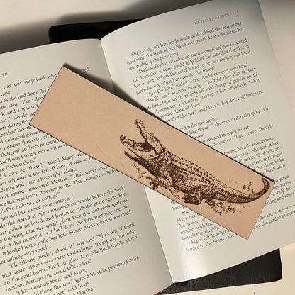 A leather bookmark with the art of an alligator on it. The leather bookmark is resting on the pages of an open book.