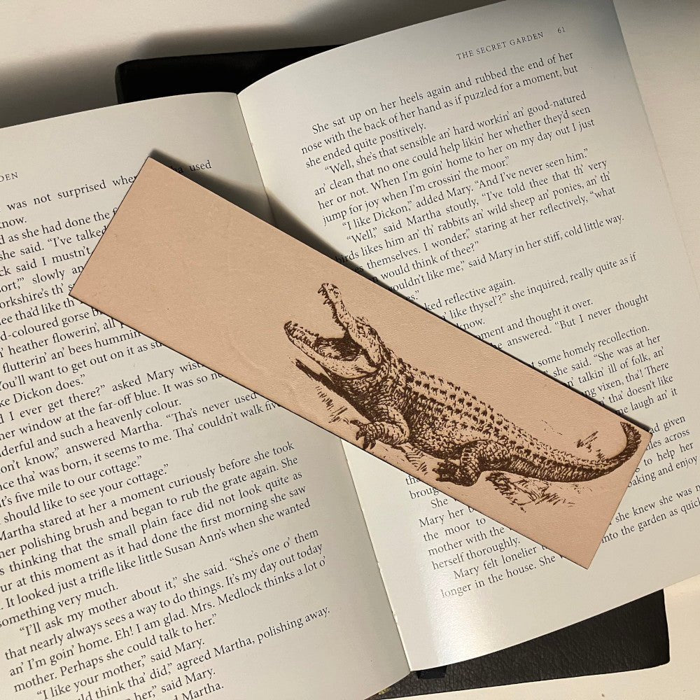 A leather bookmark with the art of an alligator on it. The leather bookmark is resting on the pages of an open book. Alligator Bookmark - Leather Bookmarks Raven King Crafts