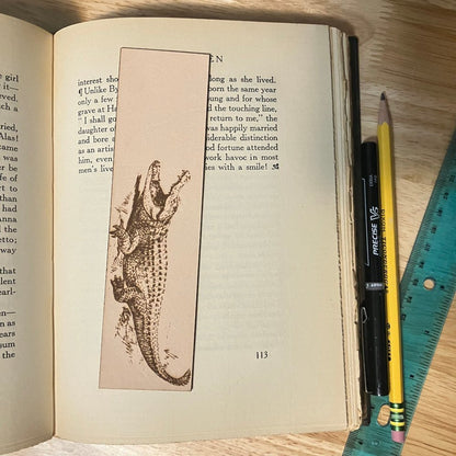 A leather bookmark with the art of an alligator on it. The leather bookmark is resting on the pages of an open book.