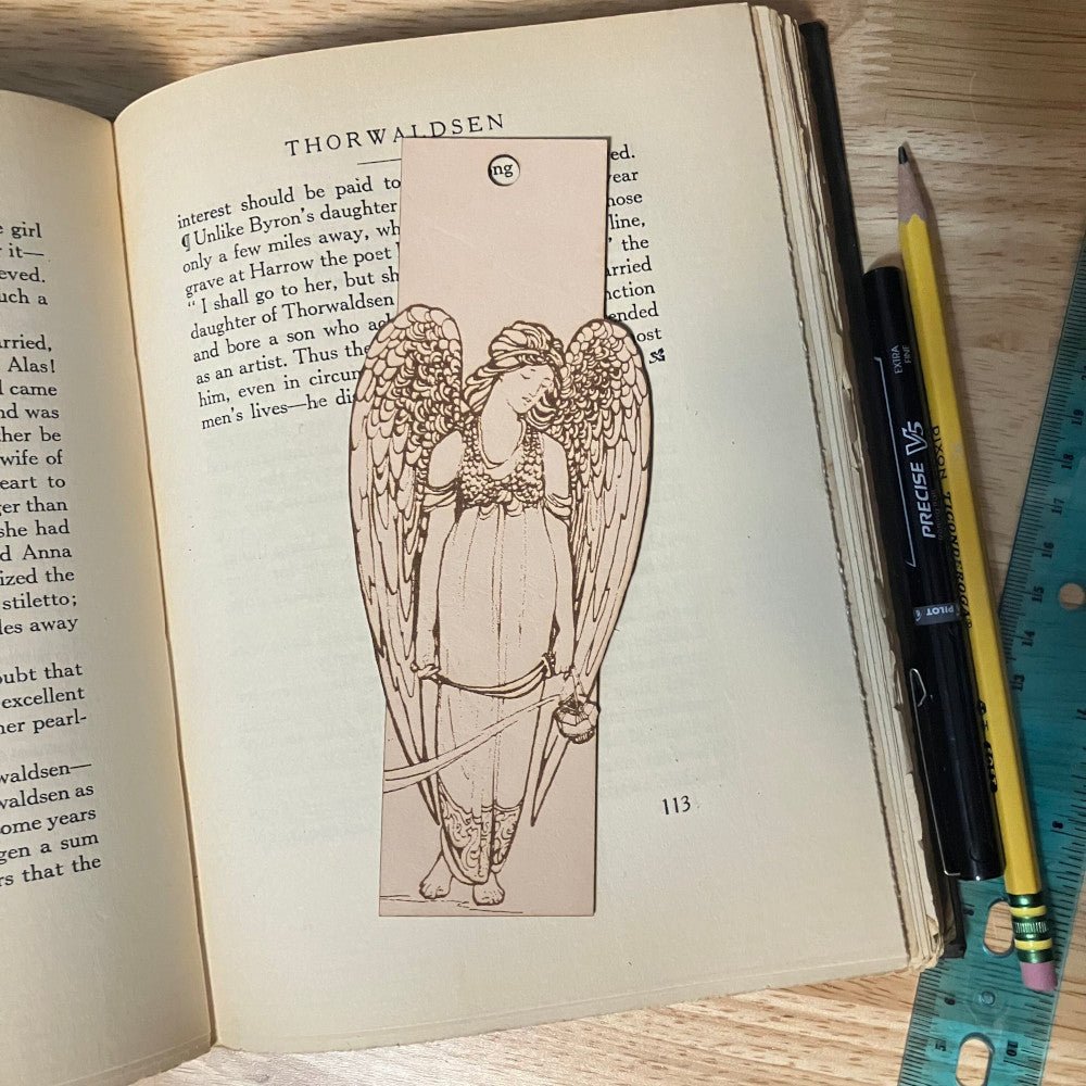 A leather bookmark of an angel. The angel is wearing a long robe. The bookmark is on the pages of an open book.