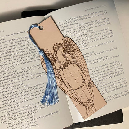 A leather bookmark of an angel. The angel is holding incense. The bookmark is resting on the pages of an open book.