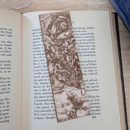 A leather bookmark with the antique art of angels and demons in battle engraved on it. The angels battle demons bookmark is resting on the pages of an open book. Angels battling Demons Bookmarks Raven King Crafts
