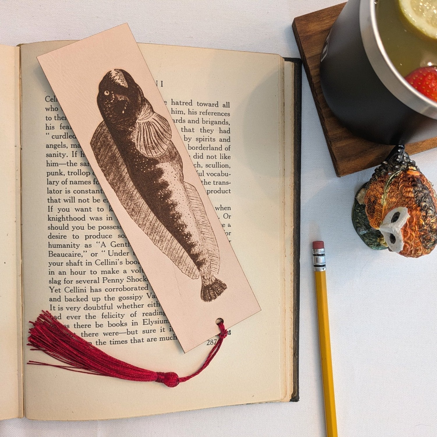 The bookmark resting on the pages of an open book. The rectangular bookmark has the antique art of a atlantic wolffish engraved on it. The bookmark also has a wine red tassel. Atlantic Wolffish Bookmark - Leather Bookmarks Raven King Crafts