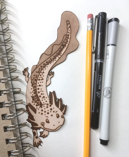 A leather bookmark shaped like an axolotl salamander. Next to it is a sketchbook and some drawing supplies for scale.