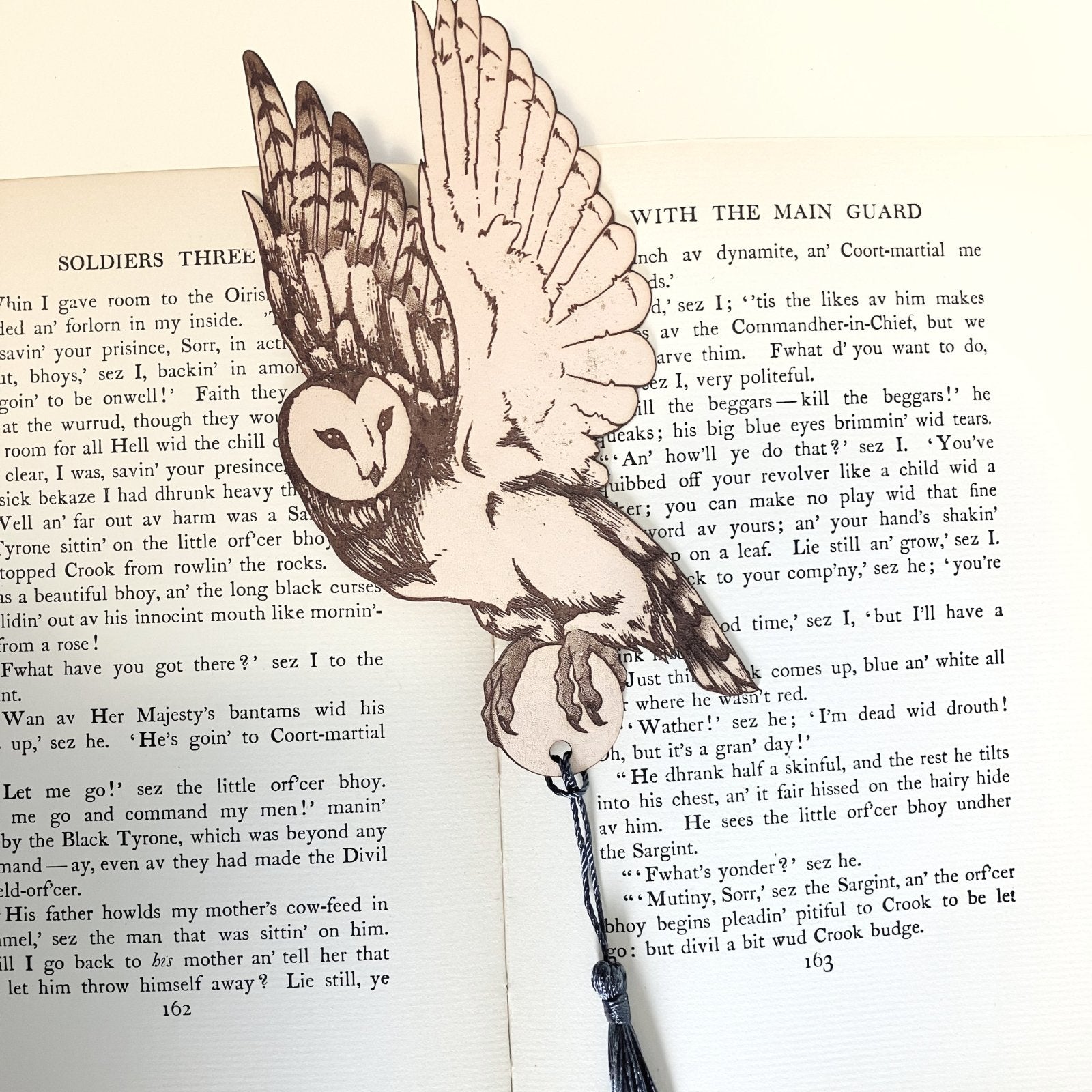 A leather barn owl bookmark, with a blue grey tassel resting on the pages of an open book.