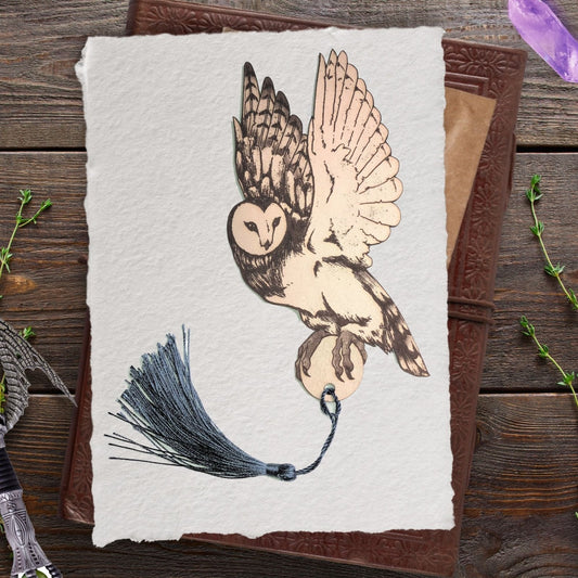 The barn owl bookmark with a tassel. It is resting a piece of paper, resting on an old leather book. The book is resting on a rustic table top.