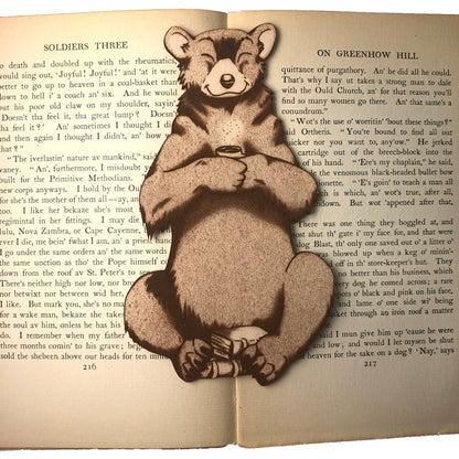 The bear with a mug, with books in its lap. The bookmark is resting on the pages of an open book.