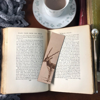 The leather black buck antelope bookmark resting on the open pages of a book. Next to the book is a pen and above is a cup.