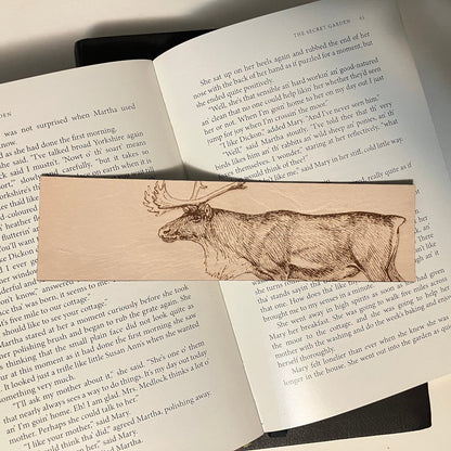A leather bookmark with the art of a caribou engraved on it. The bookmark is resting on the pages of an open book. Caribou (Reindeer) Bookmark - Leather Bookmarks Raven King Crafts