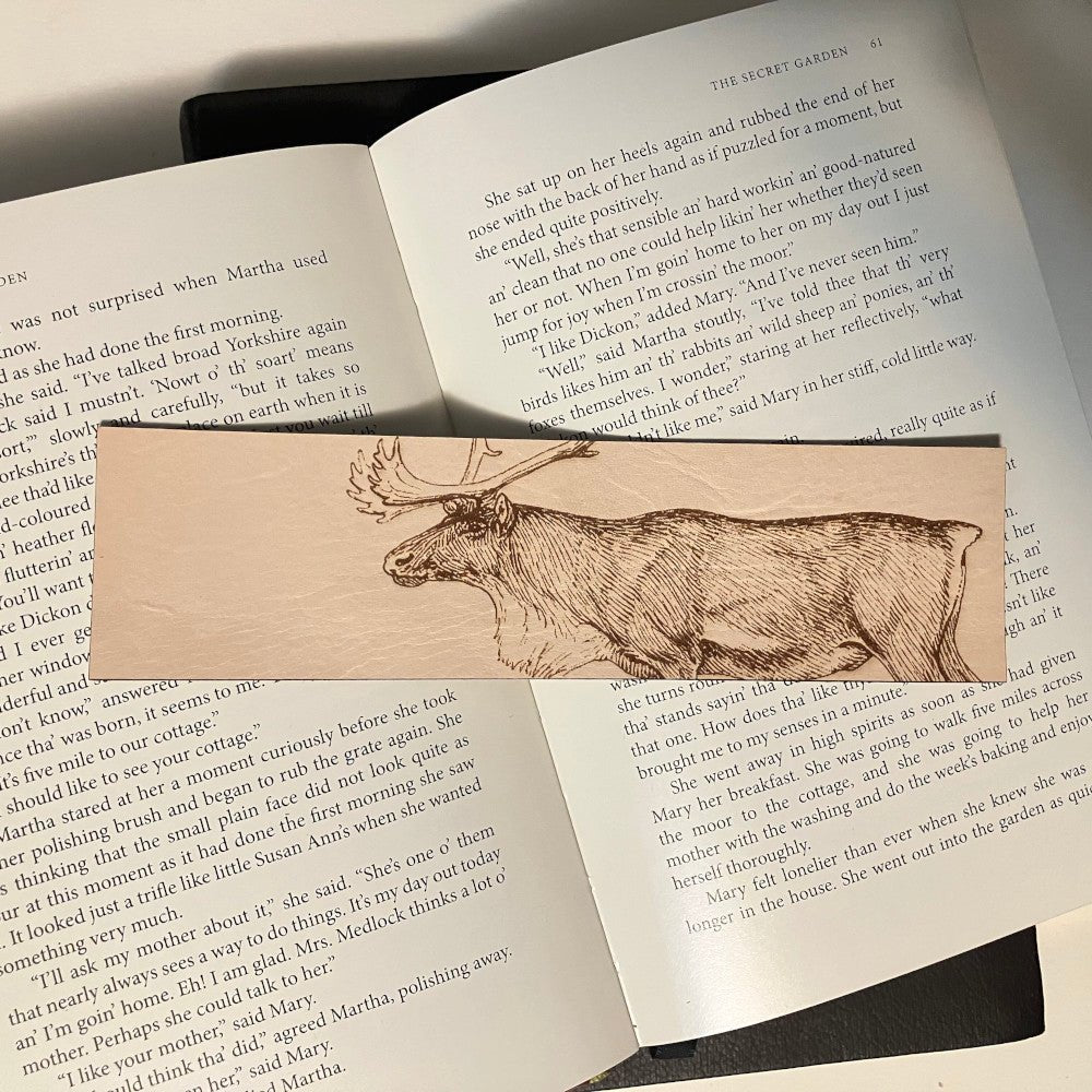 A leather bookmark with the art of a caribou engraved on it. The bookmark is resting on the pages of an open book.