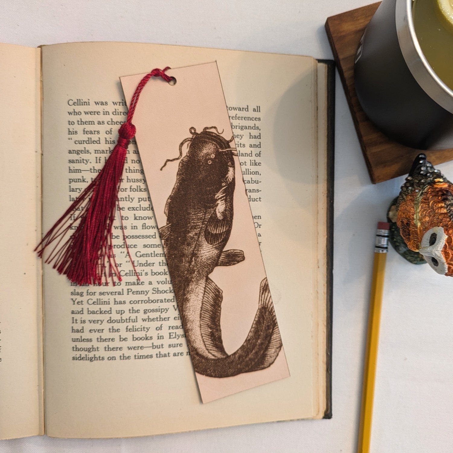 A rectangular bookmark with the antique art of a catfish engraved on it. The leather bookmark has a red tassel. Catfish (Sheatfish) Bookmark - Leather Bookmarks Raven King Crafts