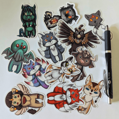 Chibi Mythos Sticker Set 1, features mythos chibis Cthulhu, kitsune, inugami, church grimm, Nekomata, spinx, Thunderbird, coyote the trickster, minotaur and Kelpie. Next to a pen for scale. Chibi Mythos Sticker Set 1 stickers Raven King Crafts
