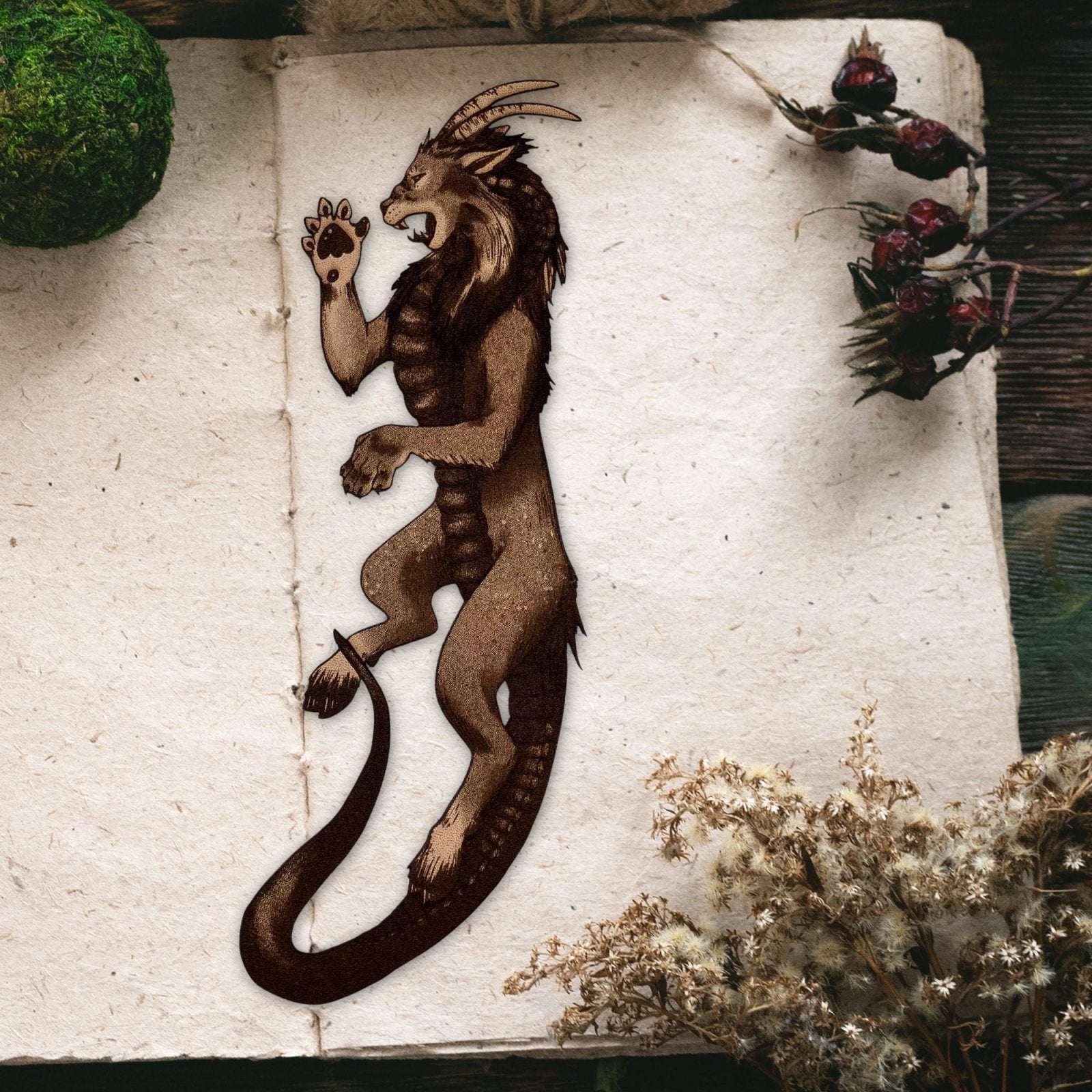 The image shows the chimera bookmark resting on the page of a an open book. The leather is cut in the shape of a chimera and engraved with the art. This is not a traditional chimera, instead this one has the head of a lion, horns of a goat and tail of a snake. Chimera Bookmark - Leather Bookmarks Raven King Crafts