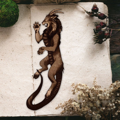 The image shows the chimera bookmark resting on the page of a an open book. The leather is cut in the shape of a chimera and engraved with the art. This is not a traditional chimera, instead this one has the head of a lion, horns of a goat and tail of a snake. Chimera Bookmark - Leather Bookmarks Raven King Crafts