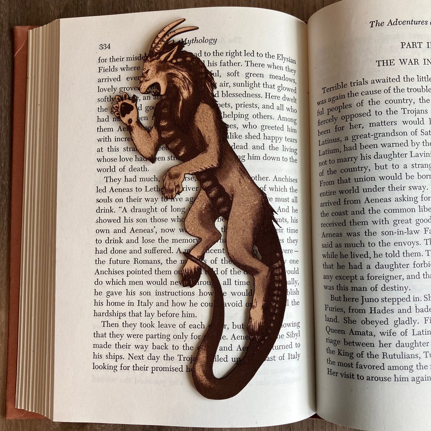 Photograph of a bookmark resting on the pages of an open book. The bookmark is shaped like a chimera. Lion like head and front paws, goat hind legs, snake tail, goat ears and horns, snake tongue. Chimera Bookmark - Leather Bookmarks Raven King Crafts