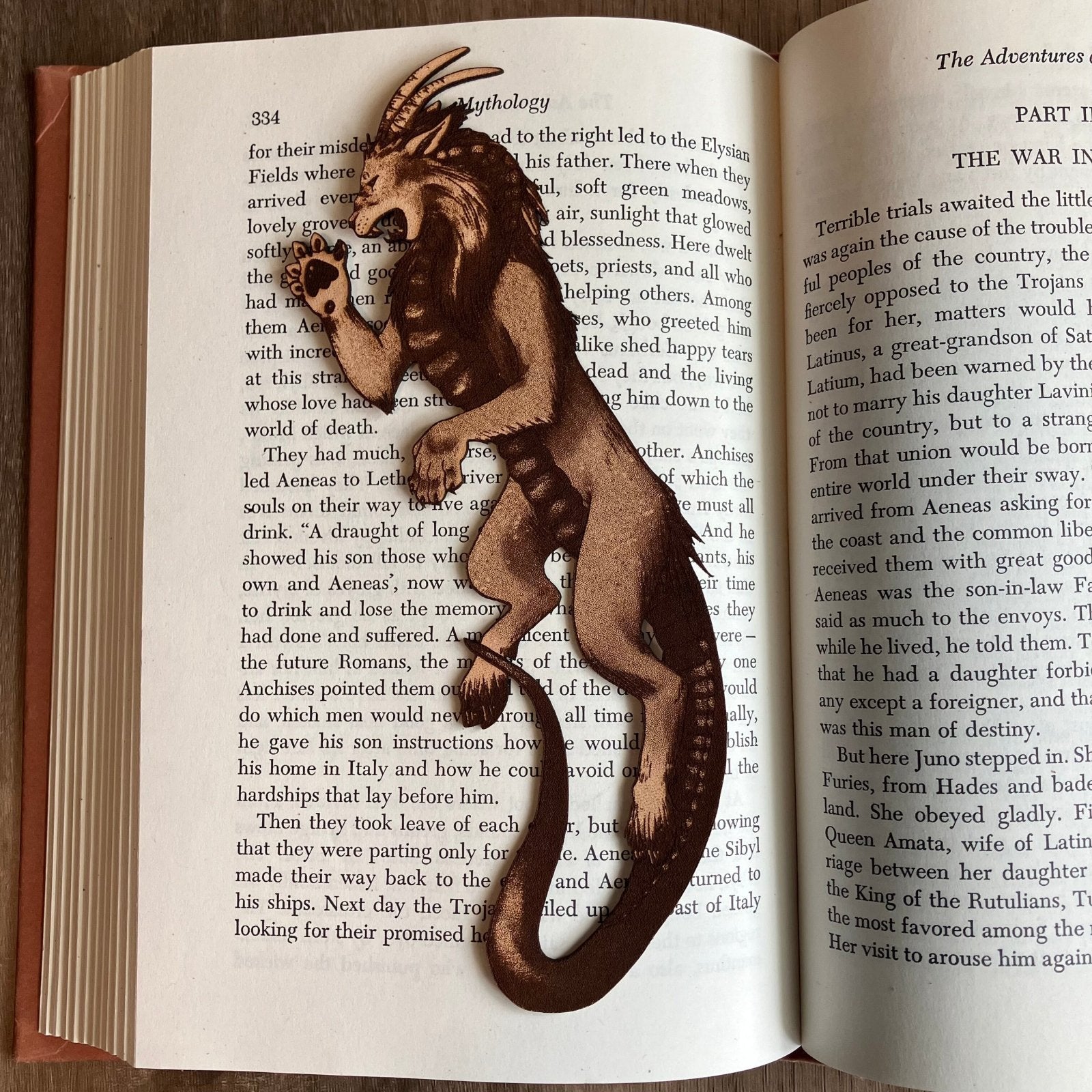 Photograph of a bookmark resting on the pages of an open book. The bookmark is shaped like a chimera. Lion like head and front paws, goat hind legs, snake tail, goat ears and horns, snake tongue. Chimera Bookmark - Leather Bookmarks Raven King Crafts