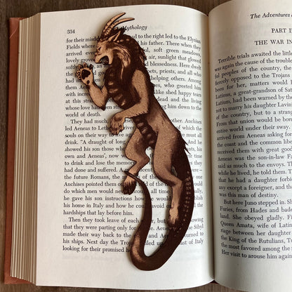 Photograph of a bookmark resting on the pages of an open book. The bookmark is shaped like a chimera. Lion like head and front paws, goat hind legs, snake tail, goat ears and horns, snake tongue. Chimera Bookmark - Leather Bookmarks Raven King Crafts