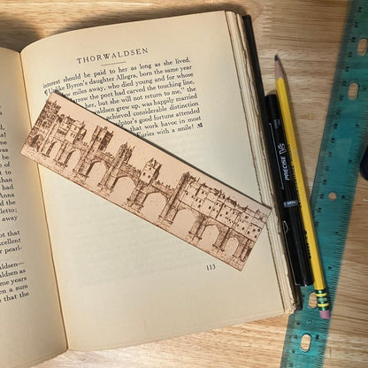 A photograph of a leather bookmark with the art of a bridge city engraved on it. The bookmark is resting on the pages of an open book. Nex to it is a ruler, pencil and pen for scale.