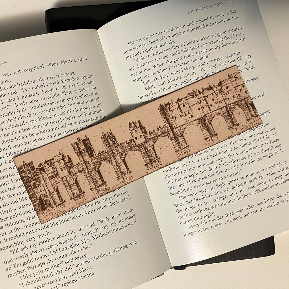 A photograph of a leather rectangular bookmark with the art of a European bridge city engraved on it. In the illustration there are buildings built onto the bridge were people used to live and work. City Bridge Historical Bookmark - Leather Bookmarks Raven King Crafts