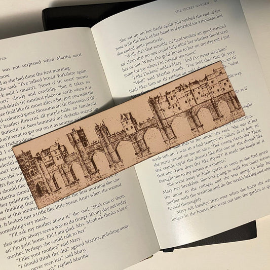 A photograph of a leather rectangular bookmark with the art of a European bridge city engraved on it. In the illustration there are buildings built onto the bridge were people used to live and work. City Bridge Historical Bookmark - Leather Bookmarks Raven King Crafts