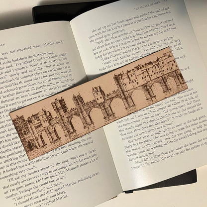 A photograph of a leather rectangular bookmark with the art of a European bridge city engraved on it. In the illustration there are buildings built onto the bridge were people used to live and work.