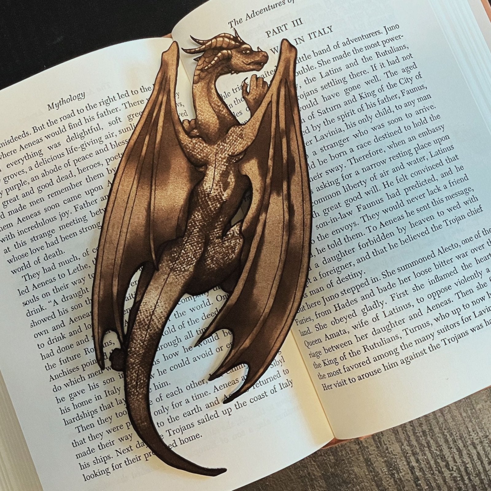 A photograph of the dragon bookmark resting on the pages of an open book. The bookmark is shaped like the a dragon. Climbing Dragon Bookmark Bookmarks Raven King Crafts