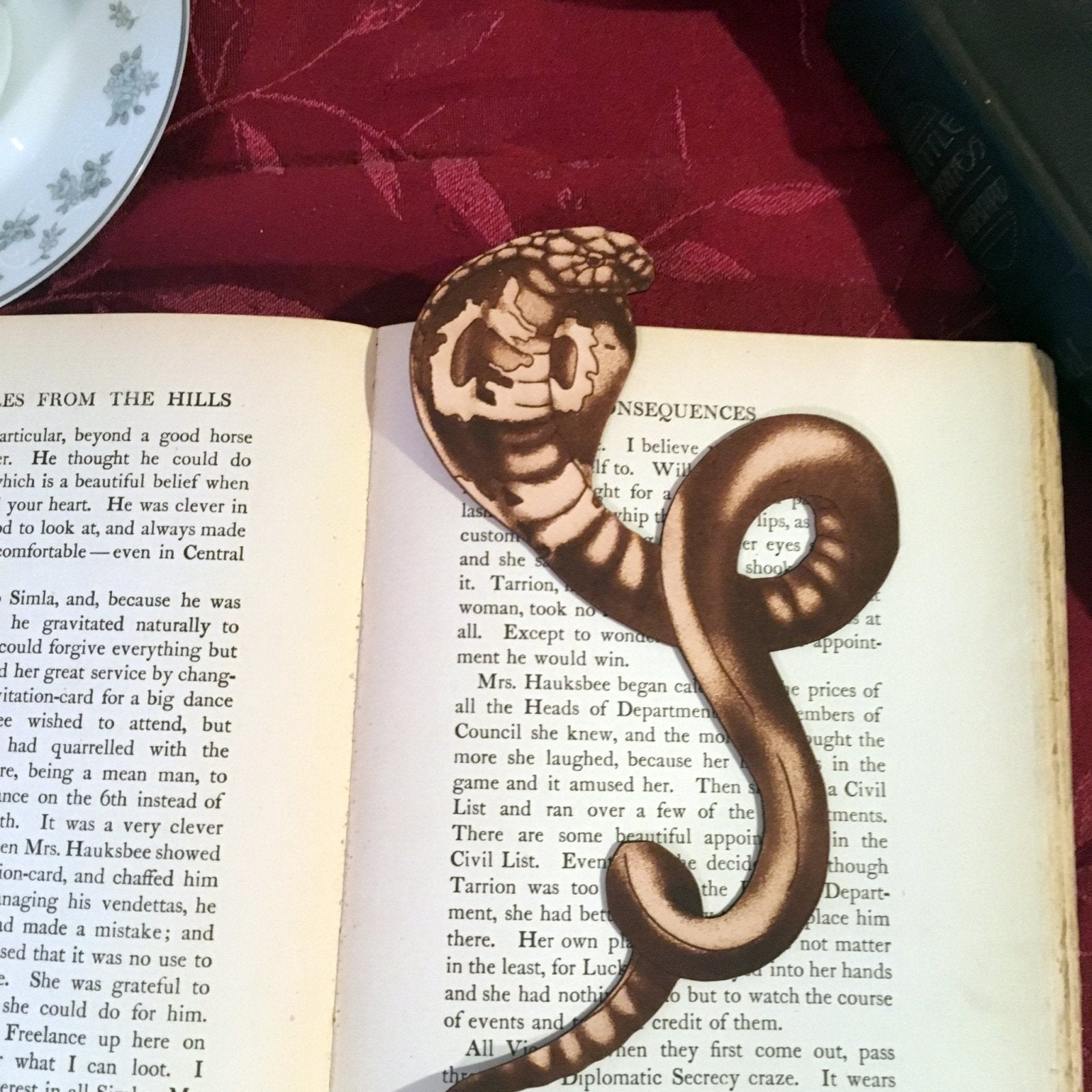 A photograph of the leather cobra bookmark. The bookmark is cut in the shape of a cobra with the art engraved on it. The snake shaped bookmark is resting on the pages of a book. 