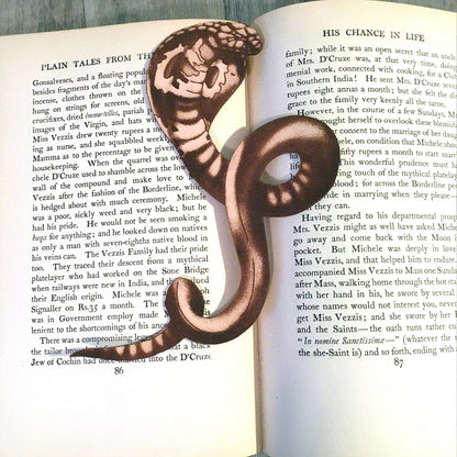 A photograph of the leather cobra bookmark. The bookmark is cut in the shape of a cobra with the art engraved on it. The snake shaped bookmark is resting on the pages of a book. 