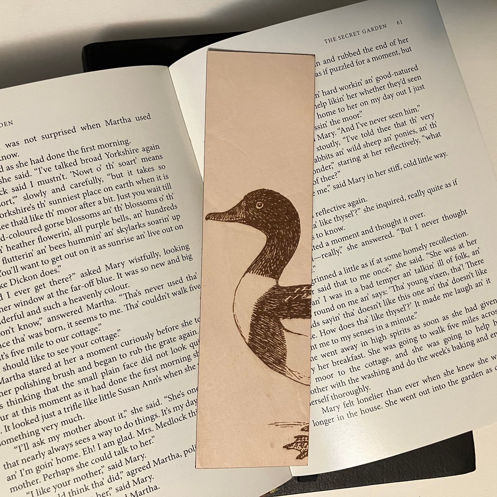 A photograph of a shelduck bookmark resting on the pages of an open book. Common Shelduck Bookmark - Leather Bookmarks Raven King Crafts