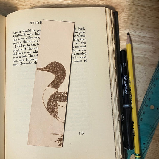 A leather rectangular bookmark with the art of a shelduck engraved on it. The bookmark is resting on the pages of an open book with a pencil to the right. Common Shelduck Bookmark - Leather Bookmarks Raven King Crafts Common Shelduck Bookmark - Leather Bookmarks Raven King Crafts