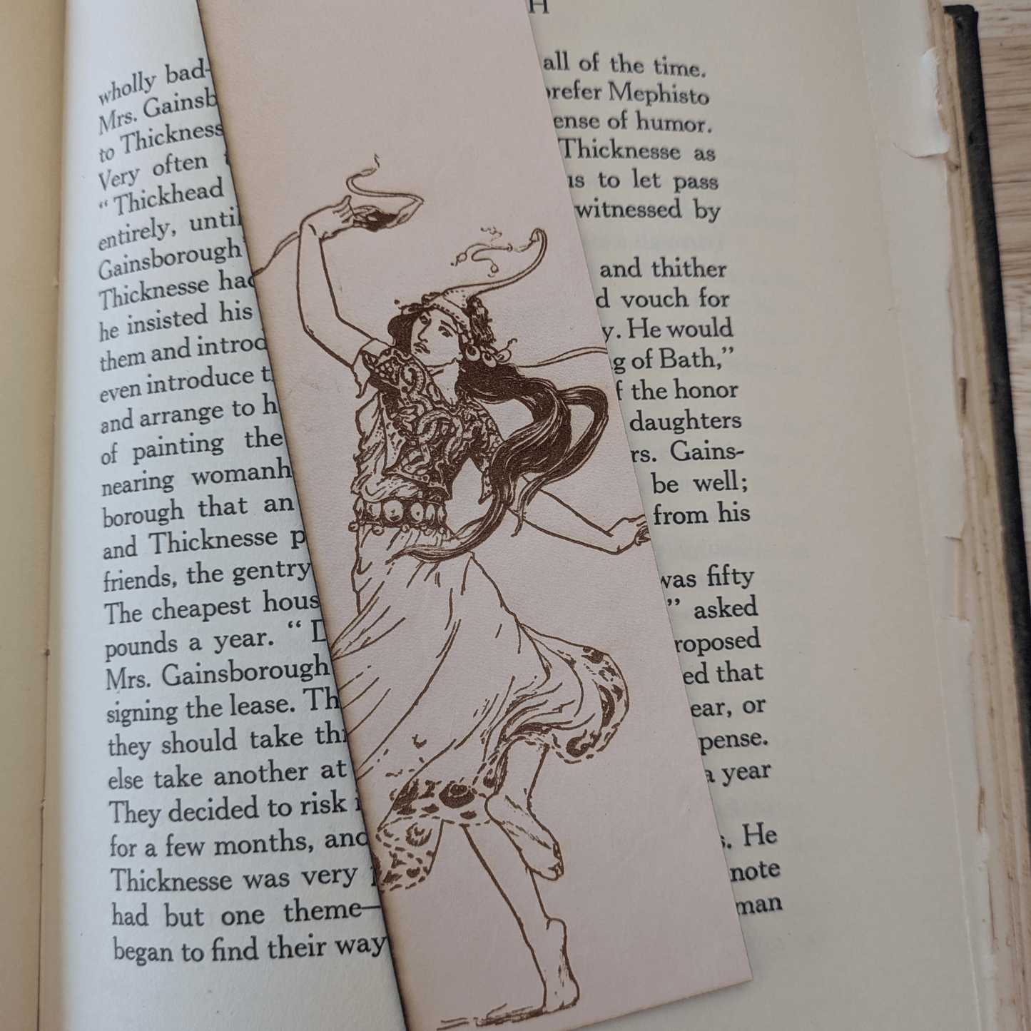 The leather bookmark with the historical art of a woman dancing engraved on it. The bookmark is resting on the pages of an open book. Dancing Woman Leather Bookmark Bookmarks Raven King Crafts