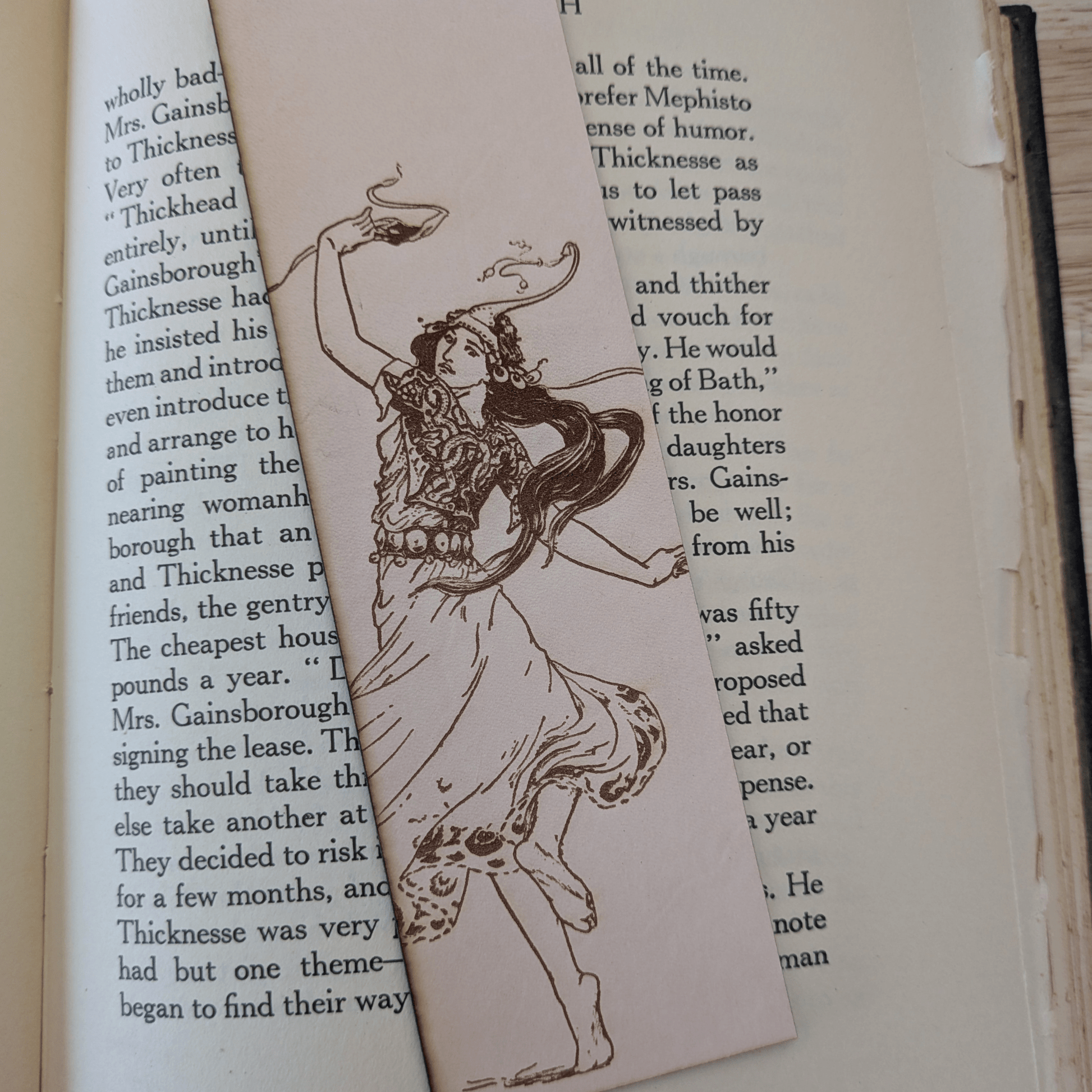 The leather bookmark with the historical art of a woman dancing engraved on it. The bookmark is resting on the pages of an open book. Dancing Woman Leather Bookmark Bookmarks Raven King Crafts
